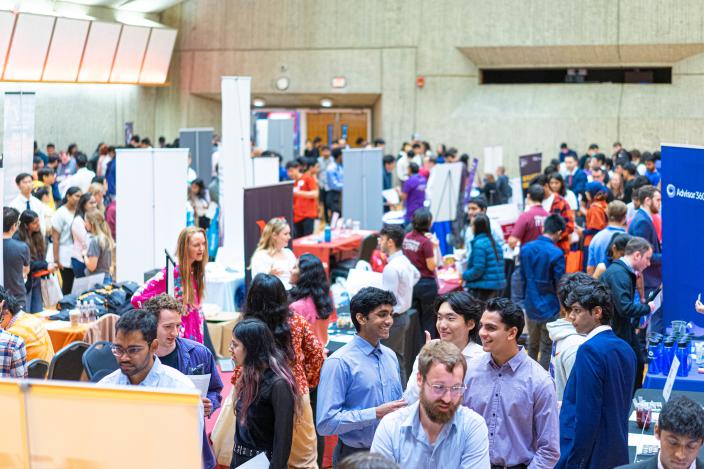 Tables of employers in the UMass Campus Center auditorium are staffed by recruiters in colorful t-shirts and surrounded by crowds of students in businesswear. 