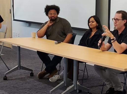 Three panelists sit at a desk and one is holding a mic