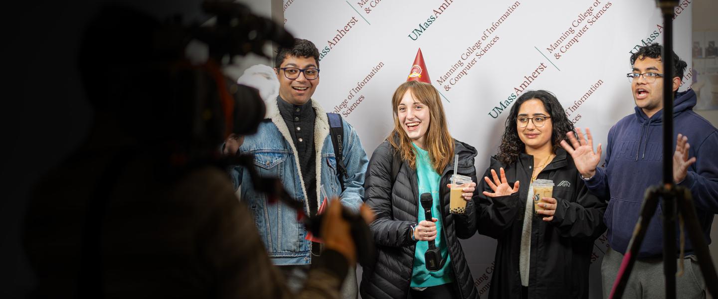 Students stand in front of a banner while a camera rolls 