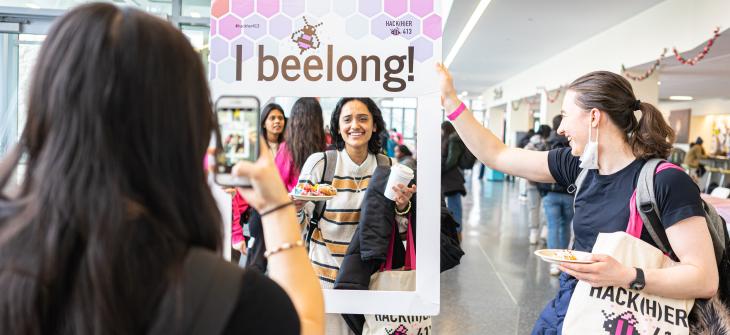 Student taking a photo of another student while holding a frame that reads: "I bee-long"